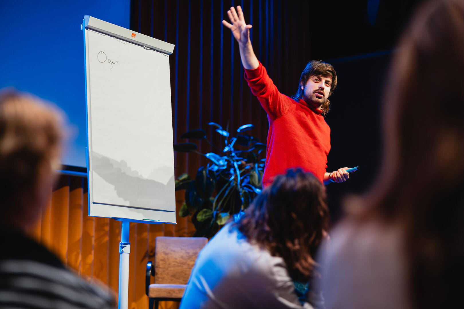 Spreker in een rood shirt houdt een presentatie voor het publiek, met een flip-over op de achtergrond.