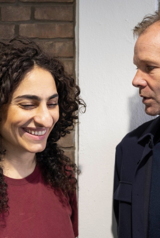 Twee mensen staan naast elkaar in een gesprek; de vrouw glimlacht terwijl de man aandachtig kijkt.