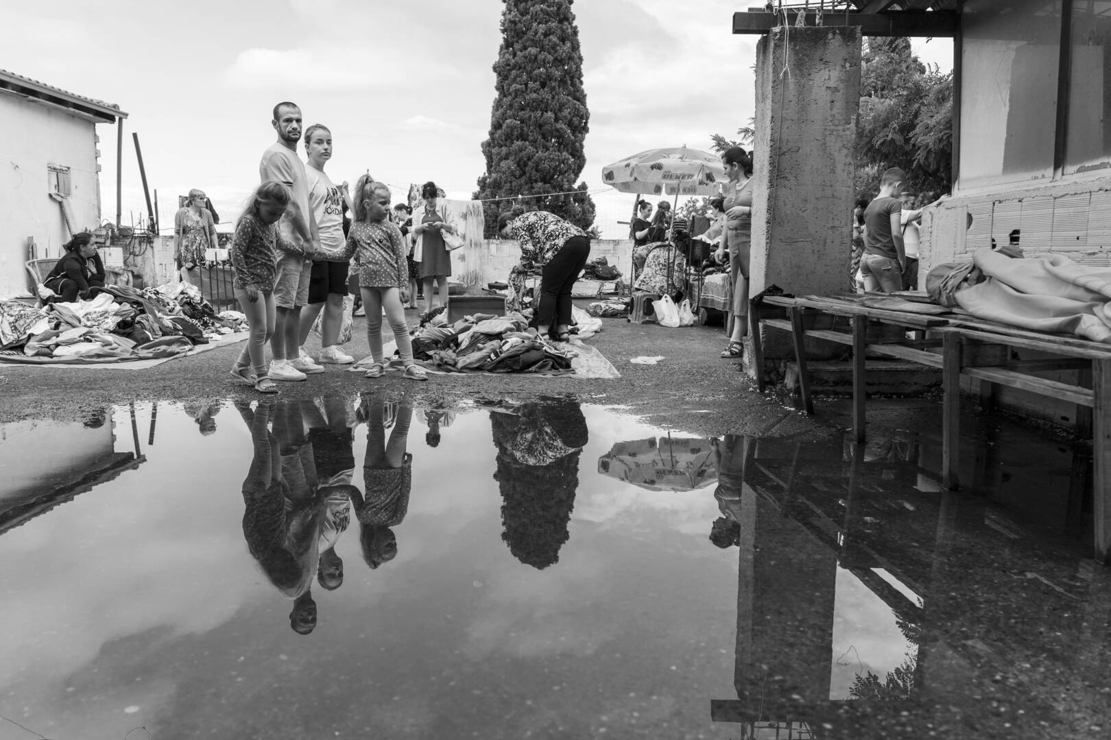 Groep mensen staat bij waterplas, met weerspiegeling, omringd door afval en gebouwen in een sombere omgeving.