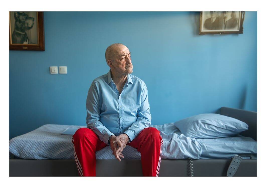 Oude man in een blauwe kamer, zittend op een bed met rode broek en glimlachend naar de horizon.